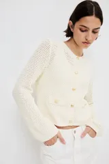 Cárdigan corto de punto texturizado color crema, con cuello redondo, cierre frontal con botones dorados y bolsillos frontales decorativos con botón dorado. Tiene terminaciones acanaladas en cuello, puños y ruedo.