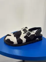 Zuecos con estampado de vaca en blanco y negro, con hebilla ajustable y suela plana.