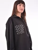 Camisa negra de corte recto, con cuello clásico, cierre frontal con botones y mangas largas con puños abotonados. Presenta dos bolsillos de parche en el pecho con apliques de tachas blancas formando un patrón geométrico.