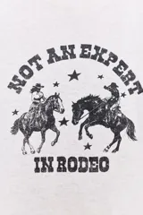 Camiseta color gris claro con cuello redondo, mangas cortas y hombros caídos. Estampado frontal con dos jinetes a caballo y la frase "Not an expert in rodeo".