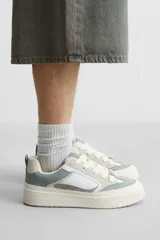 Zapatilla deportiva tipo skate con parte superior fabricada en combinación de piezas y materiales. Detalle de cordones maxi y suela con volumen. Color blanco, gris y crema.