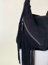 Bolso tipo hobo color tostado, hecho de cuero con flecos largos en el frente y dos bolsillos con cierre. Tiene una manija regulable con hebillas metálicas.