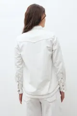 Camisa de jean color crudo, estilo western, con cuello y dos bolsillos frontales con tapa y botón. Presenta costuras contrastantes en tono rojizo.