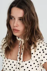 Vestido corto color crema con estampado de lunares negros. Presenta cuello redondo con lazada en la parte trasera, escote cuadrado, manga corta y cierre de cremallera oculta en la espalda.