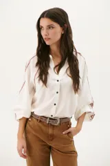 Camisa blanca de manga larga con bordados marrones en hombros y mangas, cuello en V y cierre frontal con botones.