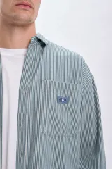 Sobrecamisa verde con rayas verticales blancas, de corte holgado, manga larga, cuello camisero y dos bolsillos frontales con solapa.