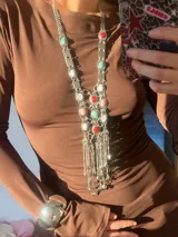 Collar largo plateado con cadena de eslabones y colgantes de piedras de colores en tonos rosa, blanco, rojo y turquesa. Incluye dijes plateados con forma de monedas y borlas de cadenas. Estilo boho.