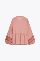 Camisa de algodón color rosa con rayas verticales finas en rojo. Tiene cuello redondo con volantes, escote pico, mangas largas con puños y bordados combinados a contraste en rojo. Cierre frontal con botones y lazada con pompones.