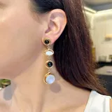 Aretes largos colgantes, con engarces de bronce, compuestos por cuatro elementos: un cabezal redondo con piedra negra, un óvalo con piedra blanca nacarada, un círculo con piedra negra y un colgante inferior redondo con piedra blanca nacarada.