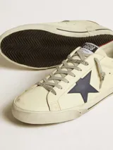 Zapatillas Golden Goose modelo Super Star, color crema con detalles en azul y rojo. Diseño vintage con efecto desgastado, estrella azul en el lateral y contrafuerte rojo.