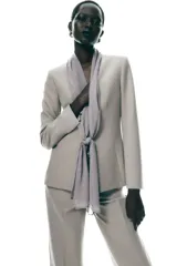 Blazer color beige con cuello solapa, manga larga con abertura y detalle de lazo gris en el frente.