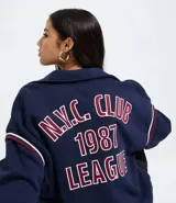 Campera estilo college de color azul marino, confeccionada en algodón con felpa, con cuello inglés, cierre delantero por cremallera y puños y bajo con rayas horizontales. Presenta estampado bordado en la espalda con el texto "N.Y.C. CLUB 1987 LEAGUE" en rojo con borde blanco.