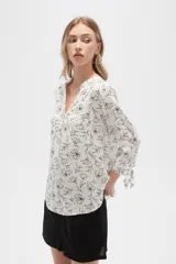 Blusa color beige con estampado de arabescos blancos, cuello mao, mangas 3/4 con lazos en los puños, cierre parcial con botones y bolsillos frontales.