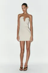 Vestido corto color crema, de escote asimétrico y sin mangas. Presenta una pieza metálica con apliques de strass en el escote.