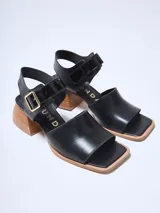 Sandalias de cuero negras con punta cuadrada, tira ancha sobre el empeine y pulsera con hebilla dorada al tobillo. Taco geométrico de madera clara.