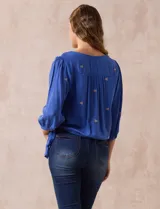 Blusa azul con bordados color crema, escote en V cruzado, mangas 3/4 abullonadas y nudo en el ruedo.