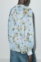 Camisa de cuello redondo y escote pico con estampado floral en tonos celeste, amarillo y negro. Manga larga abullonada acabada en puño. Detalle de volantes en el frente. Cierre frontal con botones.