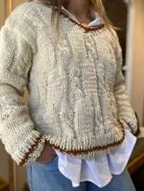 Sweater artesanal de lana color crudo con diseño de trenzas y ochos. Tiene cuello redondo y terminaciones en marrón.