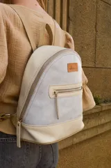 Mochila pequeña de cuero color gris claro y beige, con cierre superior y bolsillo frontal con cierre. Tiene asa de mano y correas ajustables para los hombros.