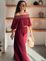 Enterito largo color blanco, con escote Bardot fruncido y mangas cortas abullonadas. La cintura es ajustable con un cinturón ancho de rafia con hebilla redonda. El pantalón es ancho y largo.