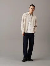 Camisa de manga larga color beige claro, con cuello clásico, cierre frontal con botones y logo de la marca bordado en el pecho.