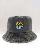 Gorro tipo bucket de denim gris oscuro con bordado frontal de la marca Rusty en amarillo y celeste.