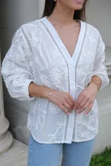 Camisa blanca con bordado de flores, cuello en V, cierre frontal con botones, mangas largas con puños elásticos y detalles de encaje.