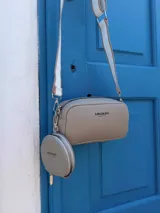 Bandolera pequeña color negro, de material símil cuero, con forma rectangular y cierre superior. Incluye un monedero redondo desmontable con cierre, ambos con logo grabado en blanco. La correa es ancha y de tela con diseño a rayas en blanco y negro.