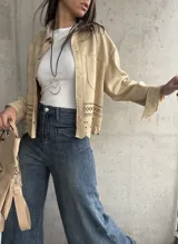 Sobrecamisa beige de gamuza con cuello camisero, cierre frontal con botones y mangas largas con puños abotonados. Presenta bolsillos de parche en el pecho y detalle de calado en el ruedo.