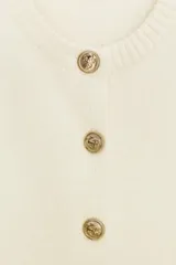 Chaleco de punto color crema, con cuello redondo y sin mangas. Cierre frontal con botones dorados.