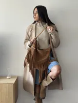 Bolso tipo hobo color tostado, hecho de cuero con flecos largos en el frente y dos bolsillos con cierre. Tiene una manija regulable con hebillas metálicas.
