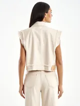 Chaqueta vaquera color crema, sin mangas, con cuello camisero, cierre frontal con botones metálicos, dos bolsillos frontales con botón y detalle de borde deshilachado.