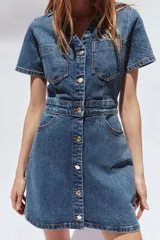 Vestido corto de denim azul, con cuello solapa, escote en pico, manga corta, bolsillos delanteros y cierre frontal con botones metálicos.