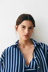 Camisa oversized de sarga a rayas verticales azules y blancas, con cuello de pico y botonadura frontal. Modelo con hombros caídos, mangas largas con tapeta y puños abotonados, y canesú en la espalda. Bajo recto con aberturas laterales redondeadas.
