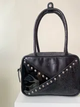 Cartera rígida negra de cuero con tachas plateadas y tres combinaciones de cuero diferentes que aportan textura. Tiene herrajes y cierre metálico plateado marca YKK. Cuenta con un asa fina para llevar al hombro o en la mano y una correa larga extra regulable.