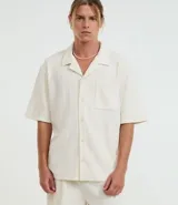 Camisa de jacquard color crema, de corte oversized, con cuello solapa, manga corta, abotonadura frontal, bolsillo en el pecho y textura en relieve.