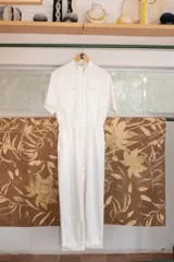 Enterito blanco tipo overol, de corte clásico con cierre frontal, pantalón largo con bolsillos, manga corta y cintura fruncida en la espalda.