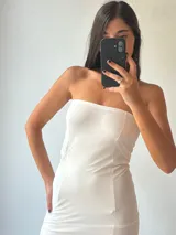 Vestido largo blanco, estilo tubo, sin tirantes (strapless). La parte superior es opaca y ajustada, mientras que la falda es de tela transparente que deja ver las piernas.