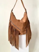 Bolso tipo hobo color tostado, hecho de cuero con flecos largos en el frente y dos bolsillos con cierre. Tiene una manija regulable con hebillas metálicas.