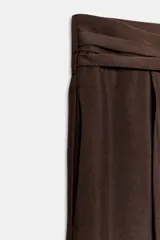 Pantalón marrón de tiro alto con bolsillos laterales ocultos en costuras, pliegues en delantero y cinturón doble en mismo tejido. Cierre frontal con cremallera y gancho metálico.