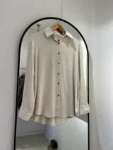 Camisa beige de lino, de corte recto y manga larga con puños arremangados. Tiene cuello camisero, cierre frontal con botones de coco y aberturas laterales.