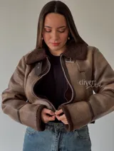 Campera corta color marrón con cuello, puños y ruedo de piel sintética. Cierre frontal con cremallera y detalles de hebillas en los laterales.