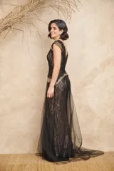 Vestido largo de tul y encaje negro con detalles dorados en los hombros y la cintura. Presenta escote en V cruzado, corte ajustado y cola de tul.