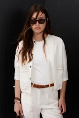 Chaqueta color crudo de lino con cuello redondo, manga tres cuartos, cierre frontal con botones y bolsillos de parche. Detalle de costuras en contraste color marrón.