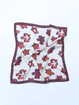 Pañuelo cuadrado de seda color crema con estampado de flores rojas, azules y blancas. Tiene borde marrón.