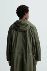Parka verde militar de tejido técnico ligero, corte oversize, cuello alto con capucha ajustable, manga larga con puños elásticos, cintura ajustable con cordón interno, bolsillos con solapa, bajo asimétrico con abertura trasera ajustable y cierre frontal con cremallera oculta y botones a presión.