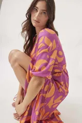 Vestido midi cruzado con estampado floral abstracto en tonos violeta y naranja, con mangas 3/4 abullonadas y lazo ajustable en la cintura. Presenta un tajo lateral.
