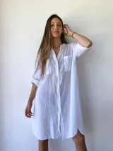 Vestido camisero blanco de corte holgado, con cuello mao, cierre frontal con botones, mangas 3/4 con puños abotonados, dos bolsillos de parche en el pecho y aberturas laterales en el ruedo.