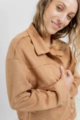 Chaqueta color beige de corte recto, estilo camisa, confeccionada en gamuza con elastano. Presenta cuello camisero, cierre frontal con botones metálicos, bolsillos grandes en el frente y charreteras en los hombros.