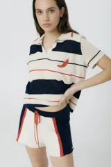 Remera polo tejida color crema con rayas horizontales en azul marino y rojo. Presenta cuello clásico, corte relajado y bordado de un ají rojo en el pecho.